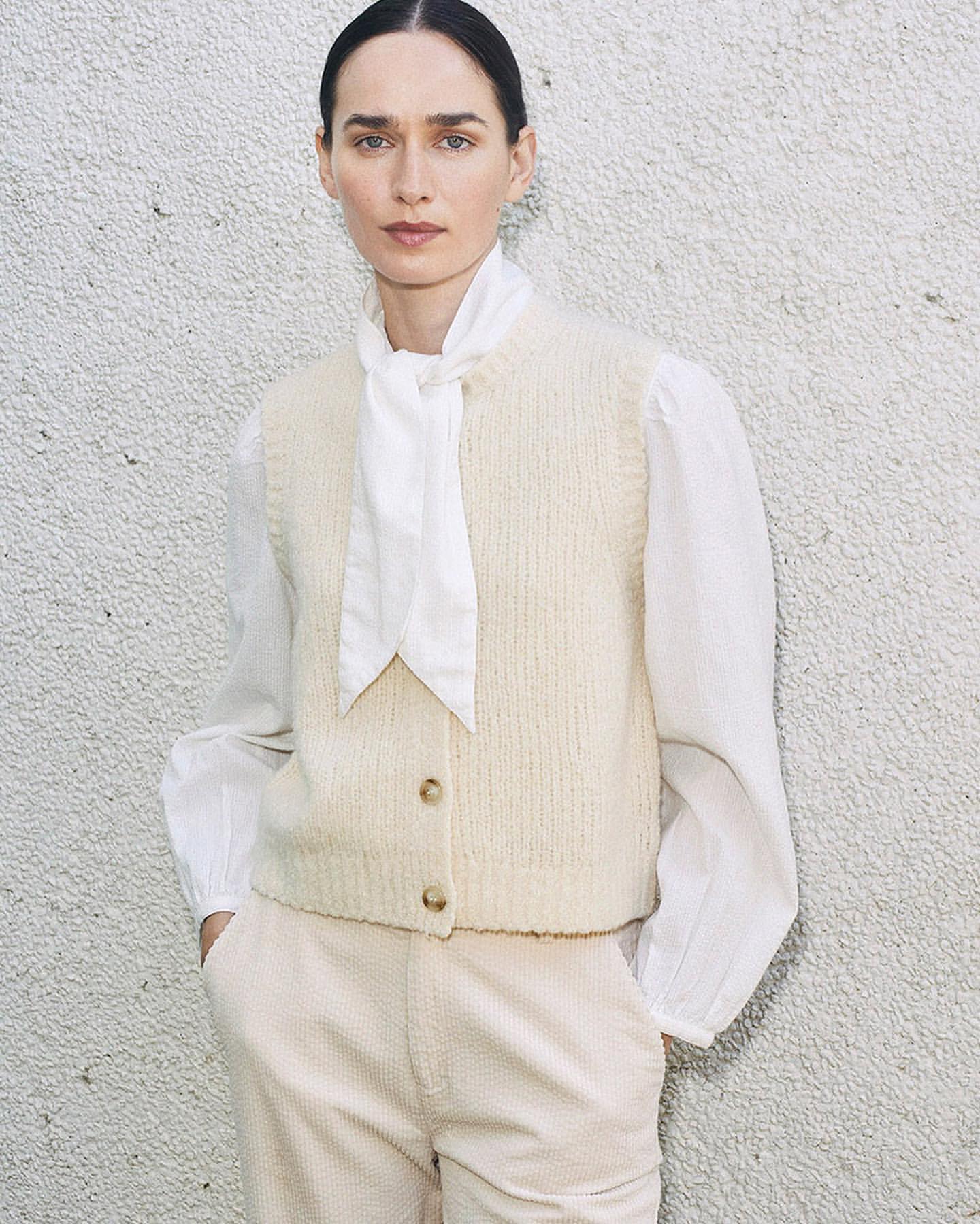 A woman poses in a cream sweater vest, white blouse with a tie, and matching pants against a textured wall, shot in natural light.