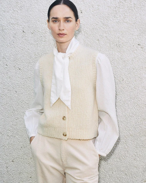 A woman poses in a cream sweater vest, white blouse with a tie, and matching pants against a textured wall, shot in natural light.