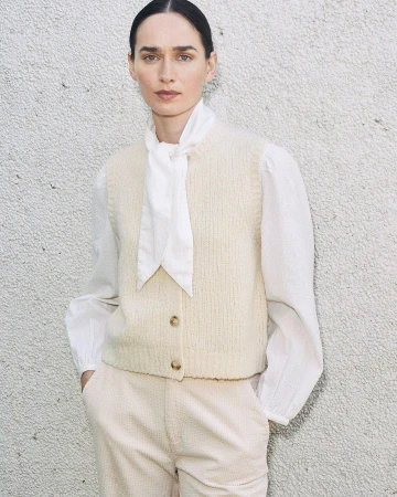 A woman poses in a cream sweater vest, white blouse with a tie, and matching pants against a textured wall, shot in natural light.