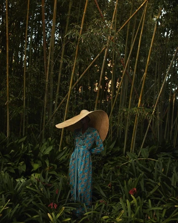 Model wearing a wide-brimmed straw hat and geometric print dress standing amidst dense bamboo stalks.