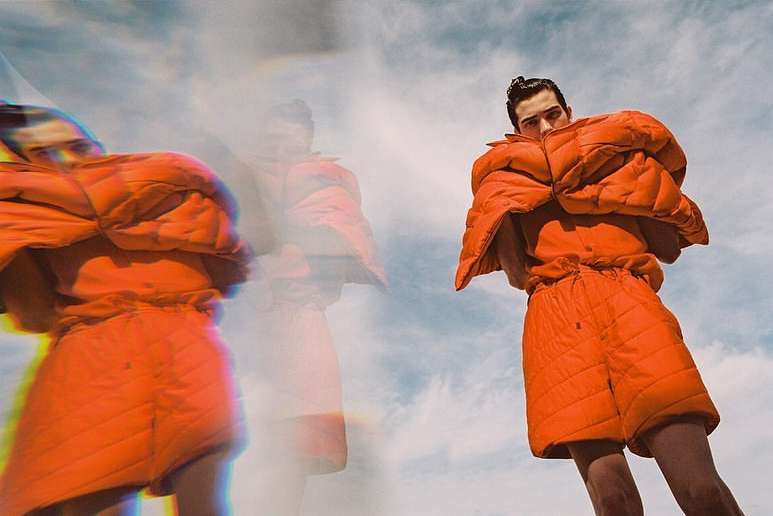 Young adult man posing in an orange quilted jacket and matching shorts against a cloudy sky backdrop, creating a fashion editorial look.