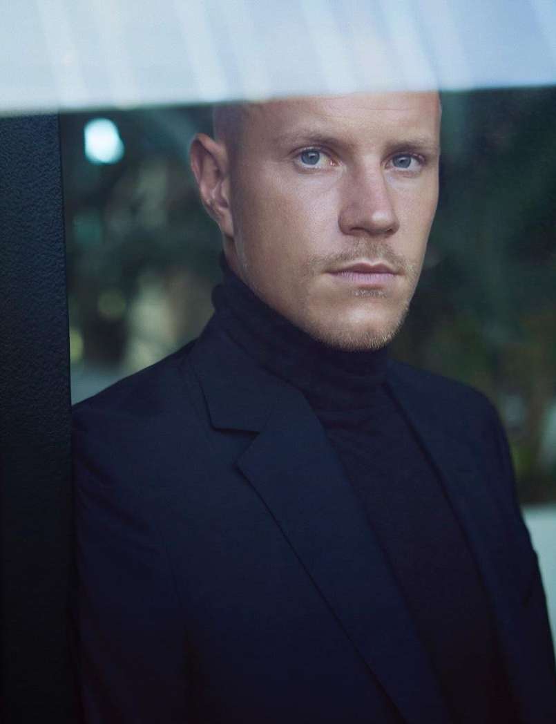 Portrait of a fair-skinned man in a black turtleneck and blazer, looking seriously at the camera through a window.