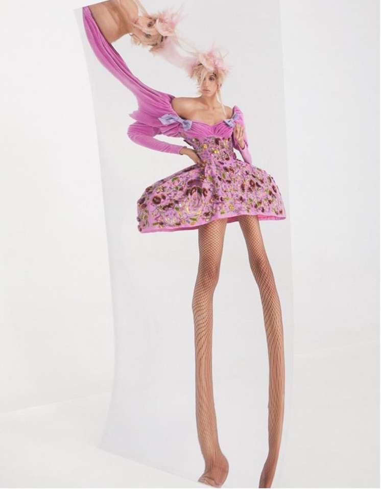 Model with exaggerated elongated legs wearing a pink patterned dress and feathered hat in a surreal vertical portrait.