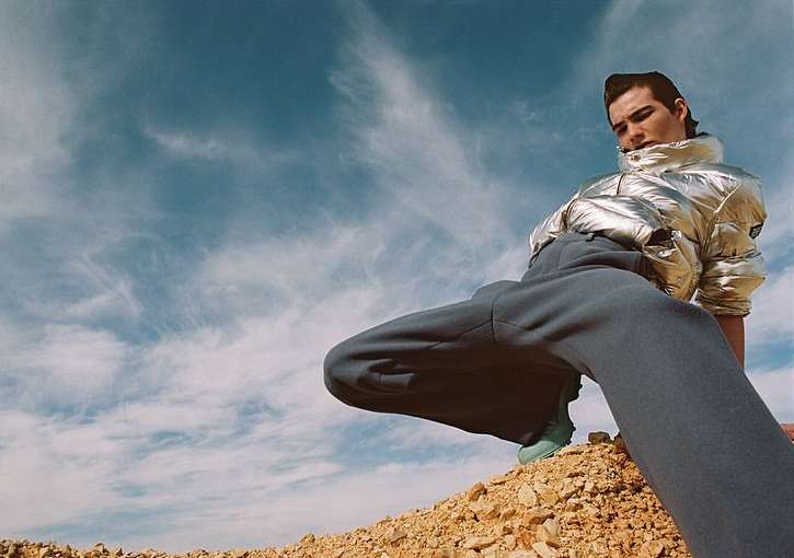 Adult male model wearing a silver puffer jacket and grey sweatpants poses on a rocky hill with a blue sky and white clouds in the background.
