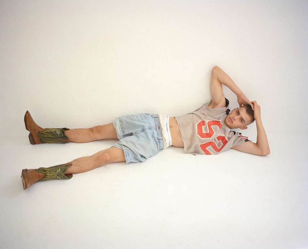 A young adult male poses lying down wearing a mesh jersey with the number 52, blue jean shorts, and brown cowboy boots on a white background.