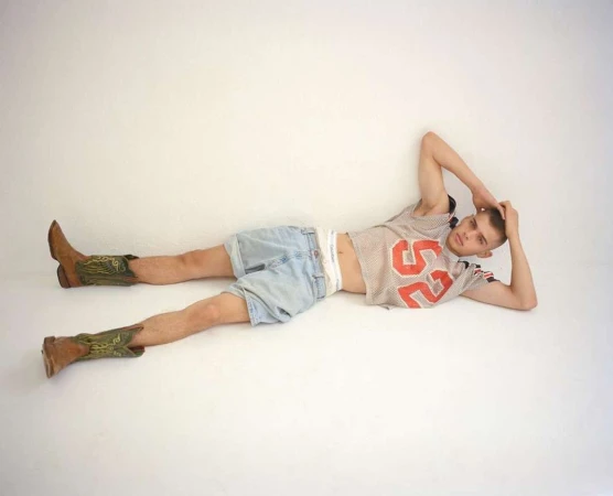 A young adult male poses lying down wearing a mesh jersey with the number 52, blue jean shorts, and brown cowboy boots on a white background.