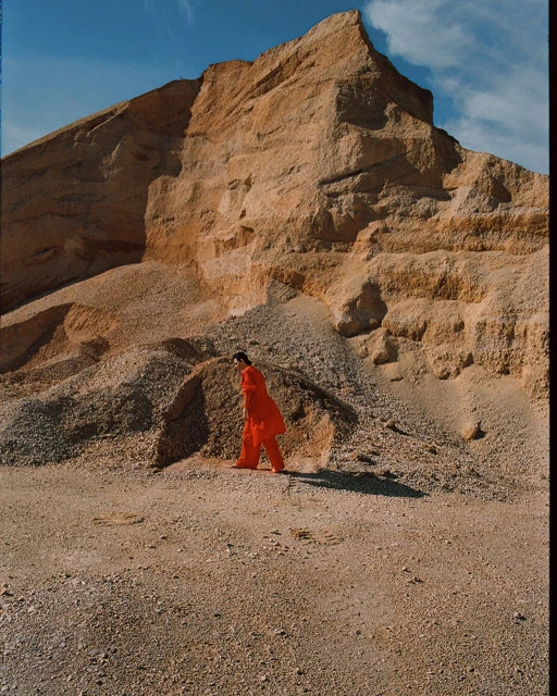 Adult woman in a red suit walking on gravel near a large earth and gravel mound against a blue sky with light white clouds.