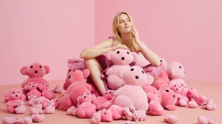 Blond woman in pink dress sits among many pink teddy bears, surrounded by pink background creating a soft and whimsical aesthetic.