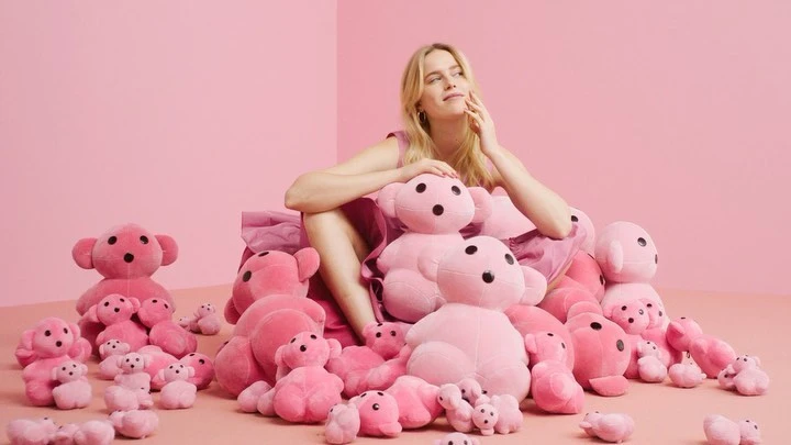 Blond woman in pink dress sits among many pink teddy bears, surrounded by pink background creating a soft and whimsical aesthetic.