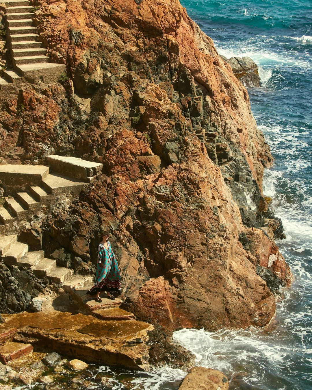 Woman in blue dress descends stone steps next to a rocky cliff and the ocean with blue water and white waves on a sunny day.