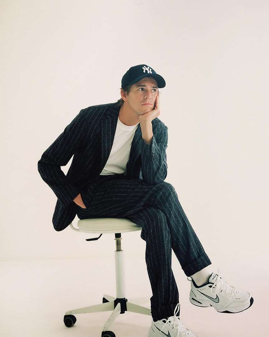 Adult male seated in a pinstripe suit and baseball cap, wearing white sneakers against a white backdrop, captured in a medium shot.