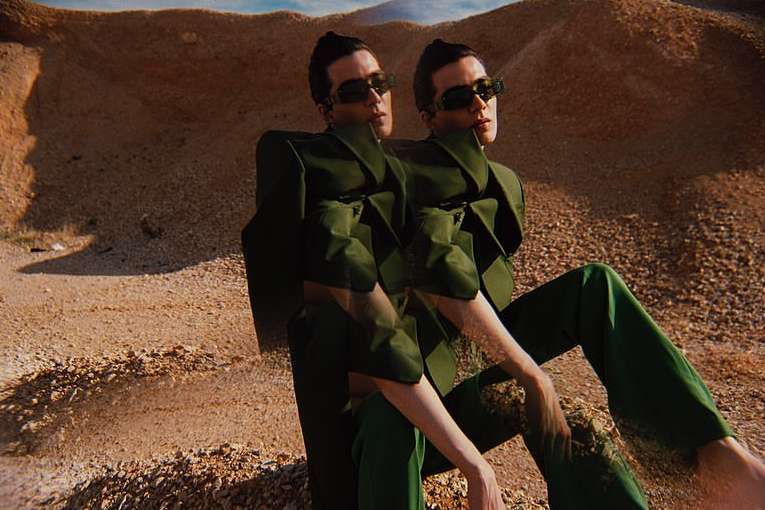 Two identical women wearing green suits and sunglasses are sitting in a desert landscape with low, sandy hills. The shot highlights fashion.