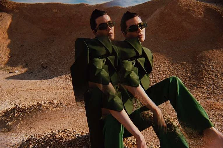 Two identical women wearing green suits and sunglasses are sitting in a desert landscape with low, sandy hills. The shot highlights fashion.