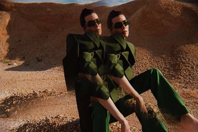 Two identical women wearing green suits and sunglasses are sitting in a desert landscape with low, sandy hills. The shot highlights fashion.