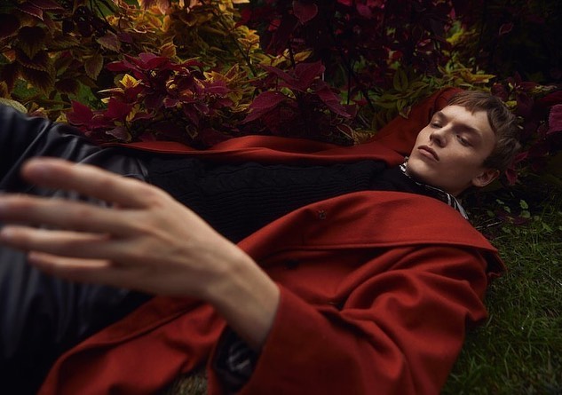 A young man in a red coat rests in foliage, creating a serene fashion editorial image with natural light and earth-tone colors.
