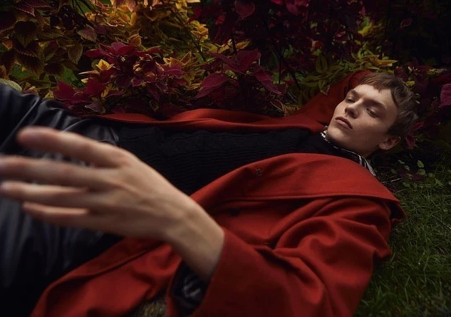 A young man in a red coat rests in foliage, creating a serene fashion editorial image with natural light and earth-tone colors.
