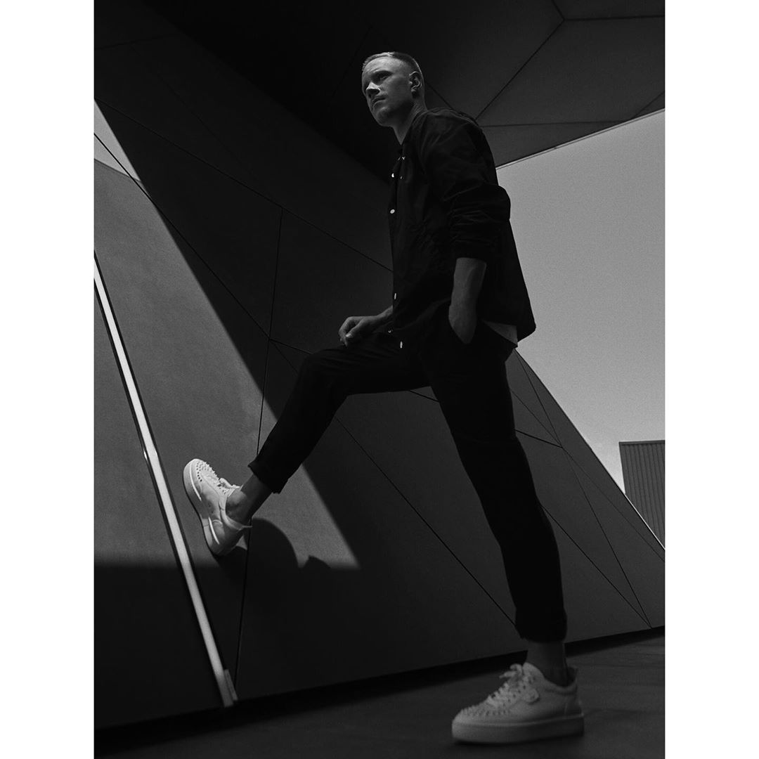 Monochrome portrait of a slender adult male model in athletic wear striking a casual pose against a geometric architecture backdrop.
