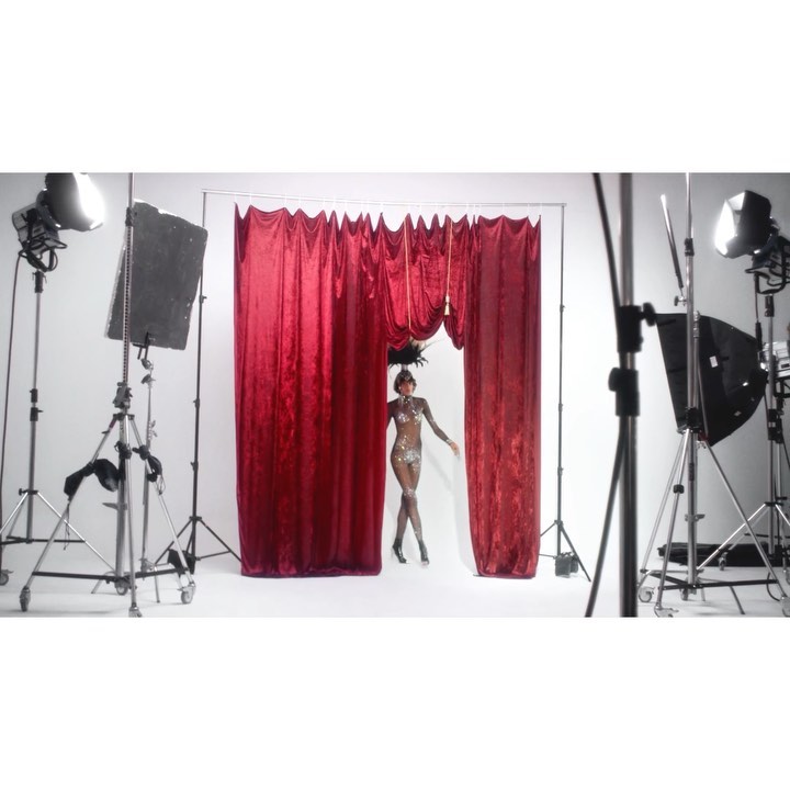A woman in a sparkling bodysuit and feathered headpiece stands between red velvet curtains in a white photo studio with lighting equipment.