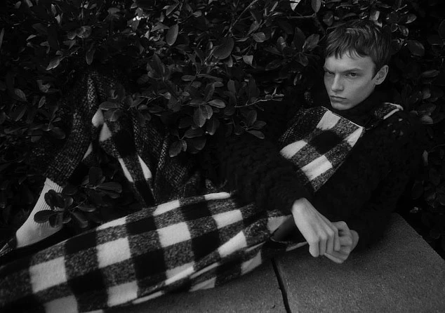 Monochrome fashion shot of a young adult male model lounging in a checkerboard suit and dark sweater, surrounded by lush green foliage.