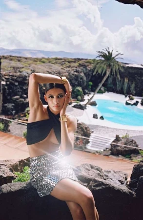 A woman in a black top and silver skirt poses outdoors with a swimming pool and volcanic landscape in the background.