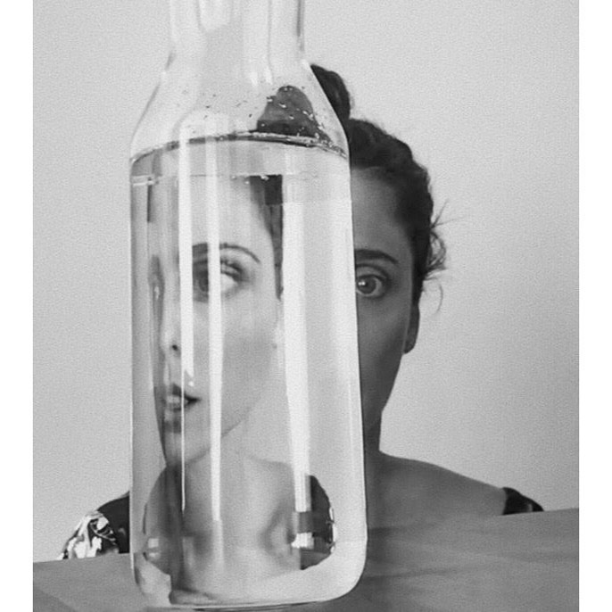 Black and white portrait of a woman partially obscured by a clear glass bottle filled with water, creating a distorted reflection of her face.