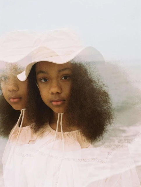 Close-up of two young African-descent children in matching light-pink dresses and hats, against a blurred beach and sky.
