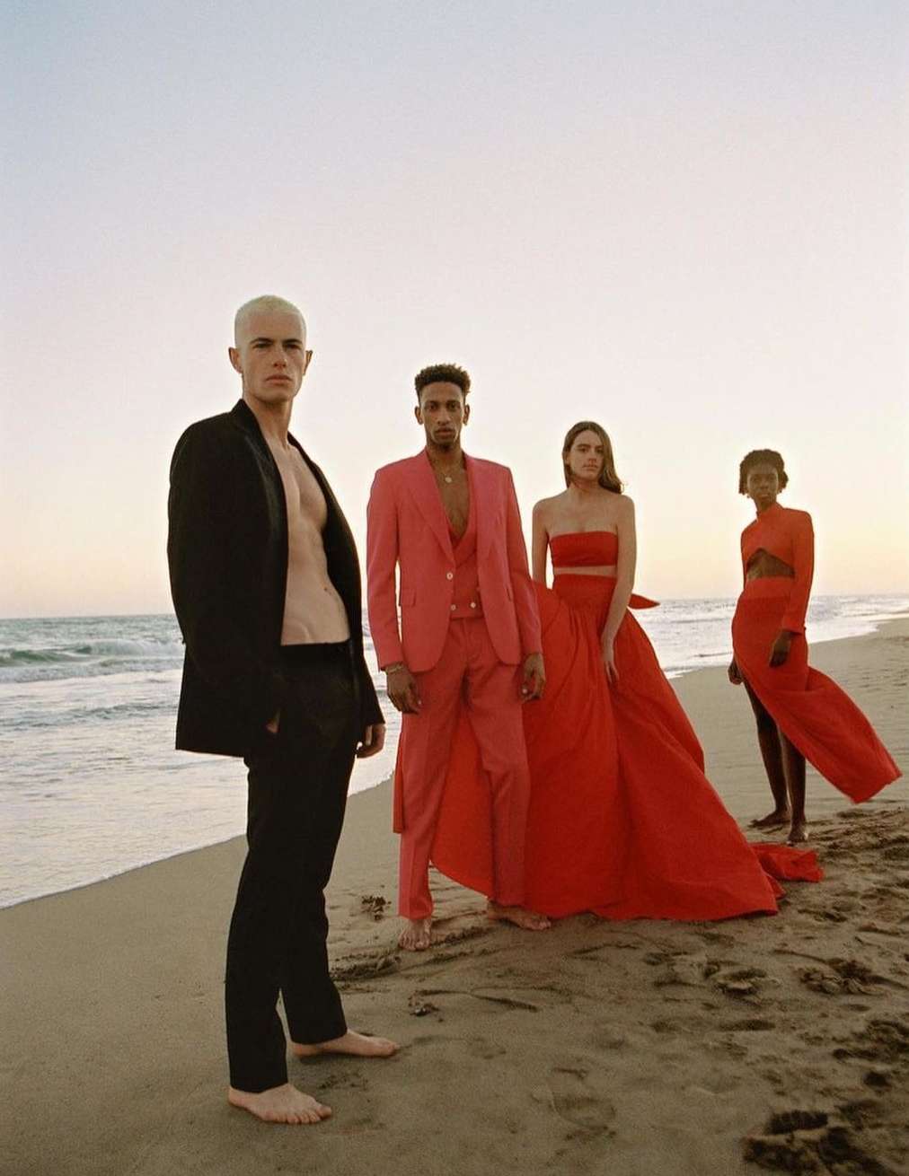 Four fashion models pose on a beach at sunset. Two women wear long red gowns; one man is in a red suit, and another is in a black suit.