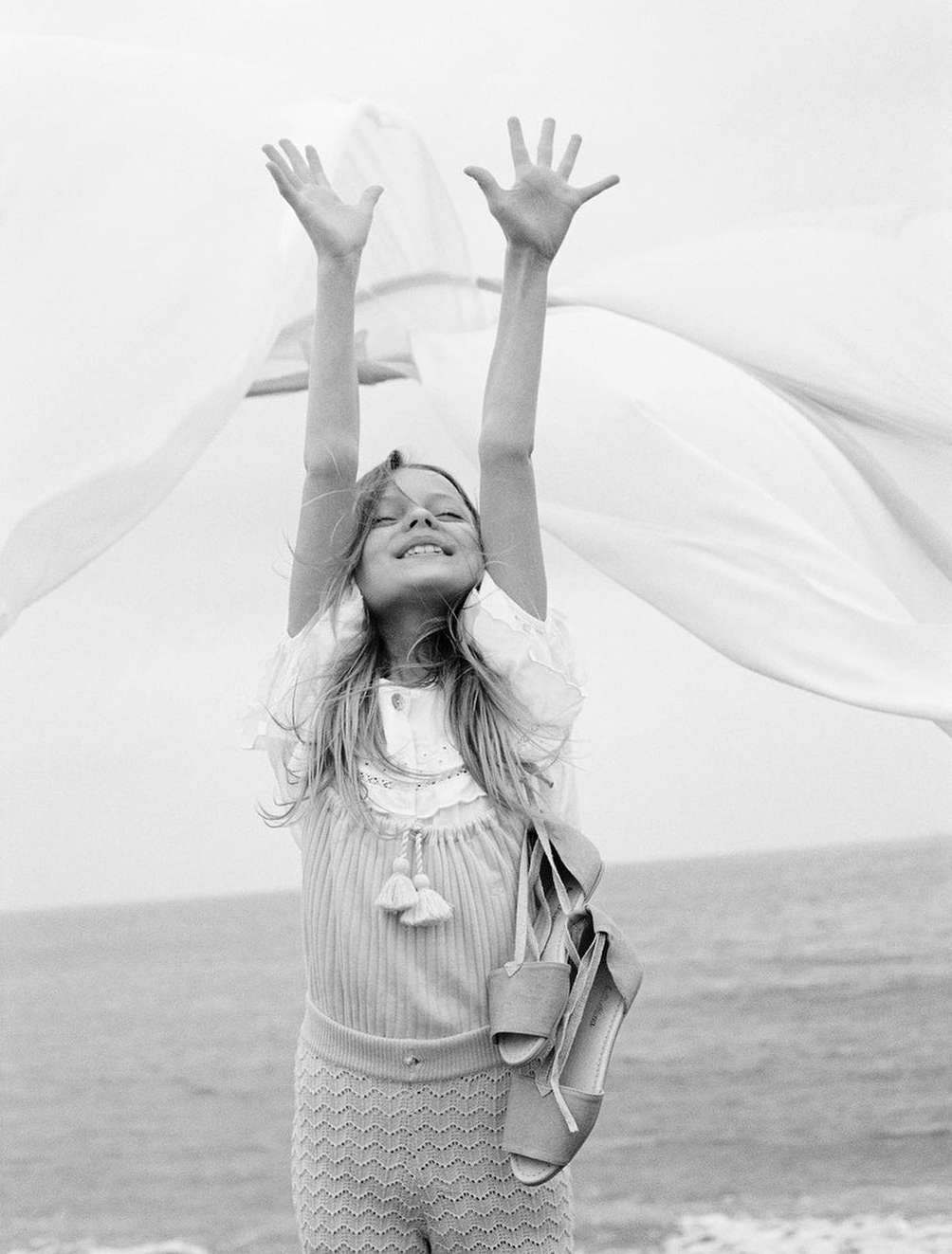 A child with arms raised stands on a beach holding a pair of sandals, with a flowing white fabric in a monochrome setting.