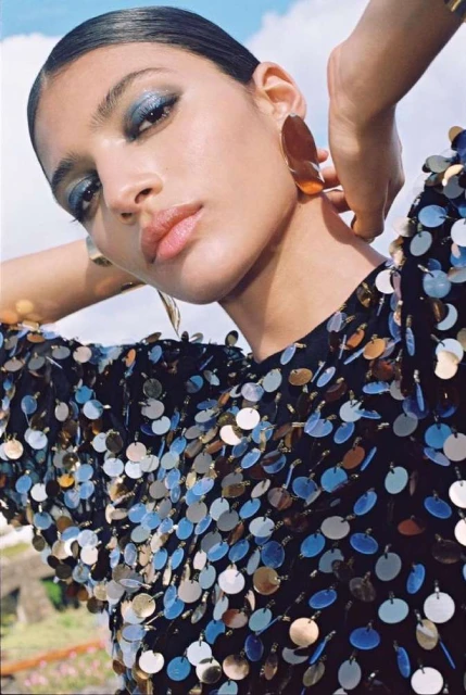 Woman with blue eyeshadow wearing a sequined top and large earrings poses outdoors with hands behind her head in a close-up shot.