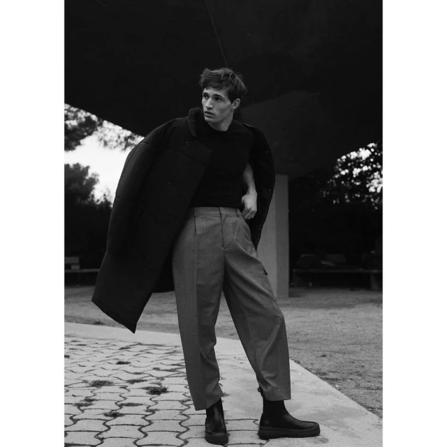 Monochrome full shot of a young adult man in a coat and casual pants standing on a patterned concrete path in an outdoor setting.