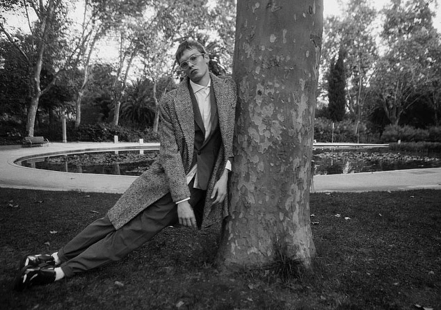 Black and white shot of a man leaning against a tree in a park, wearing a coat, suit, and glasses; pond and trees in background.