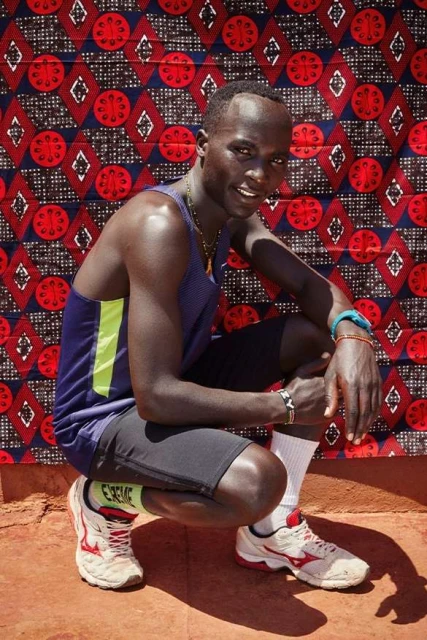 A smiling man in athletic wear crouches against a backdrop of a patterned textile. He is wearing athletic shoes, shorts, and a tank top.