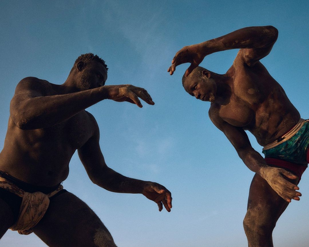 Two African male wrestlers covered in sand in a low angle outdoor shot against a blue sky, showcasing strength and tradition.
