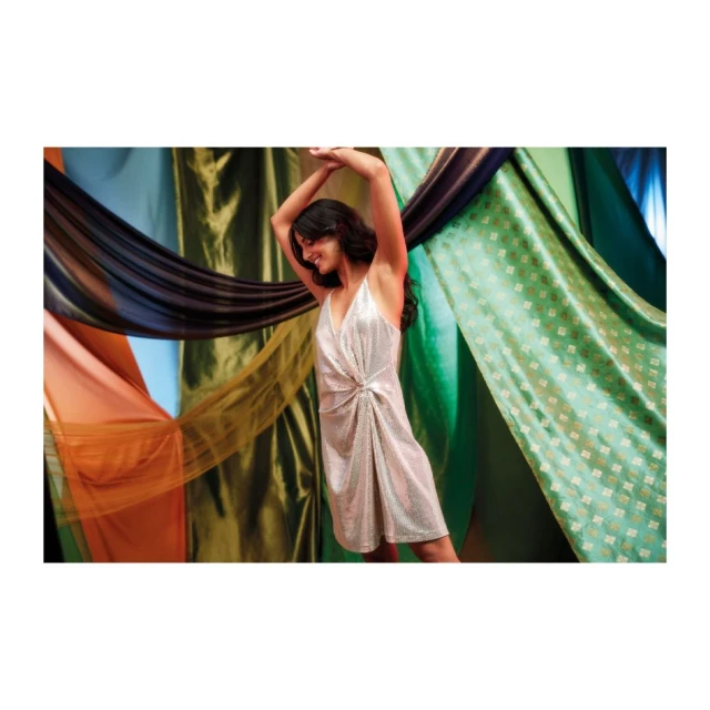 Adult woman in a sparkly dress with arms raised, smiling, against a colorful draped-fabric backdrop. Fashion, party, studio shot.