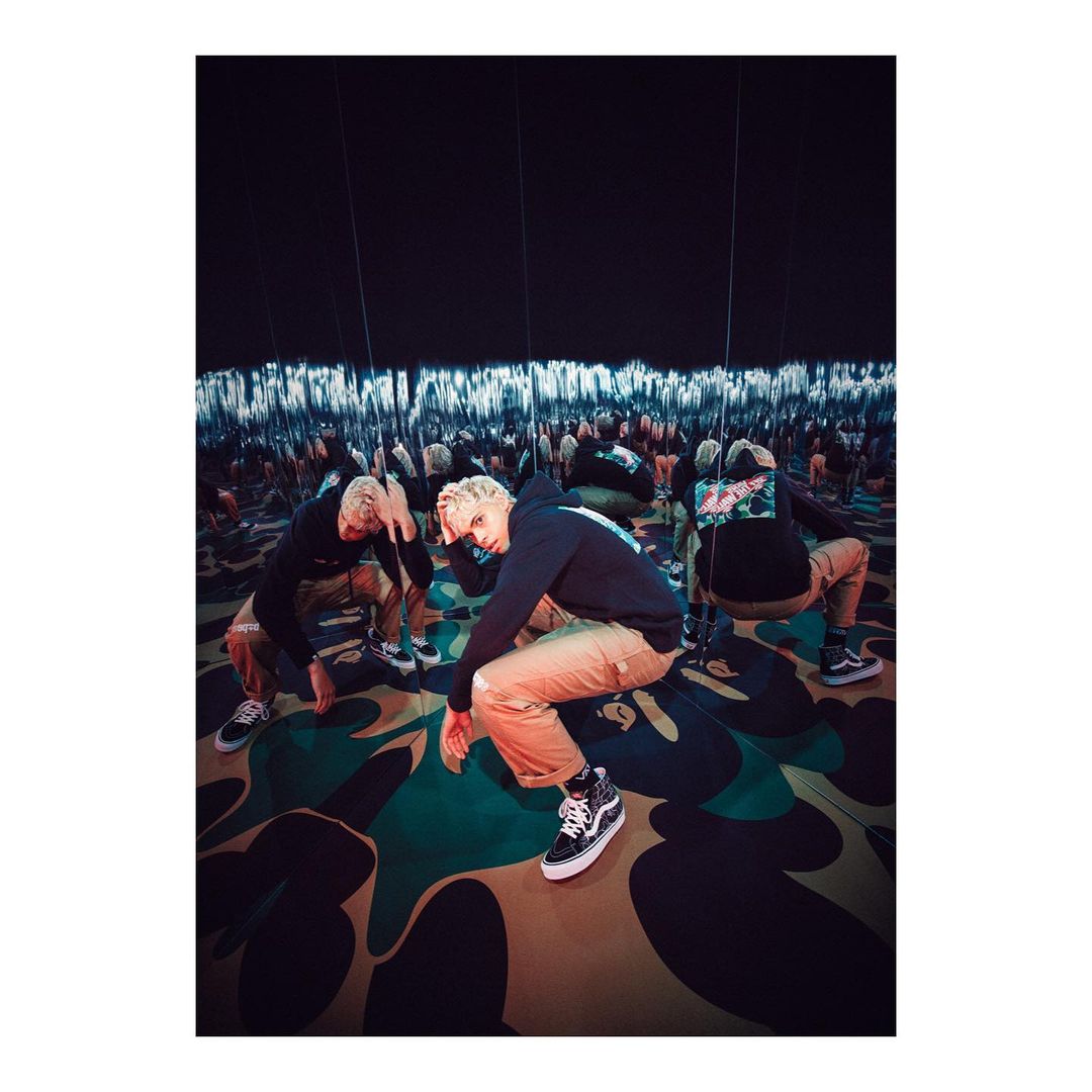 A blonde adult male squats in black sneakers and beige trousers, reflected in surrounding mirrors with patterned floor and a dark ceiling.