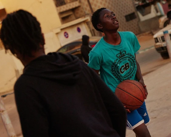 Teenager holds a basketball outdoors in daytime, wearing blue shorts and a teal t-shirt with graphic, in a youth setting.