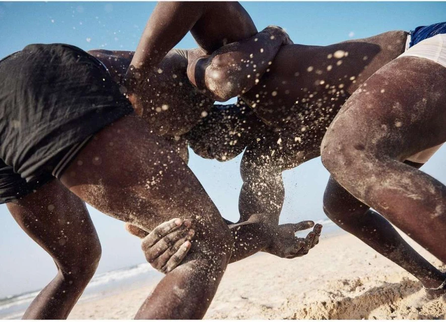 Three dark-skinned men wrestling on a beach, covered in sand, with a clear blue sky and ocean in the background. Sport and outdoor activity.