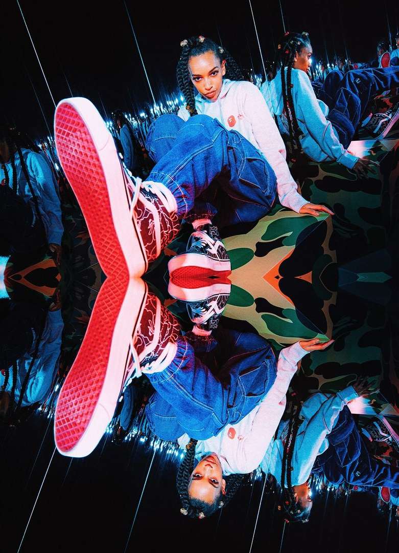 A stylish woman with braided hair sits with Vans shoes, against a reflected camouflage backdrop in cool, mirrored photo studio.