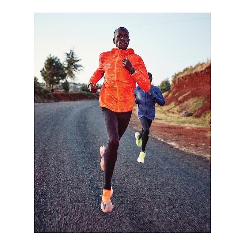 Two adult male athletes are jogging on a paved road; the first wears a bright orange jacket and the second wears a blue jacket.