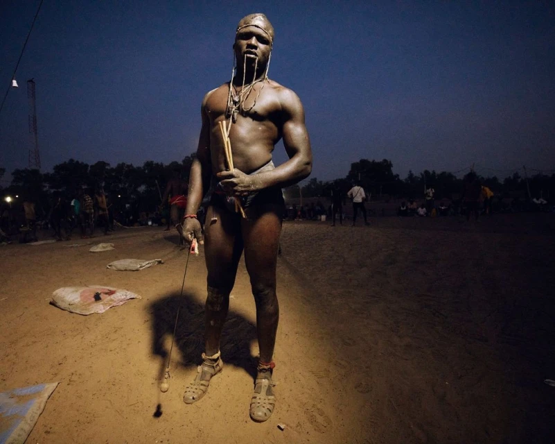 Adult male traditional wrestler standing in sandy arena, holding sticks and rope whip under twilight sky. Audience in background.