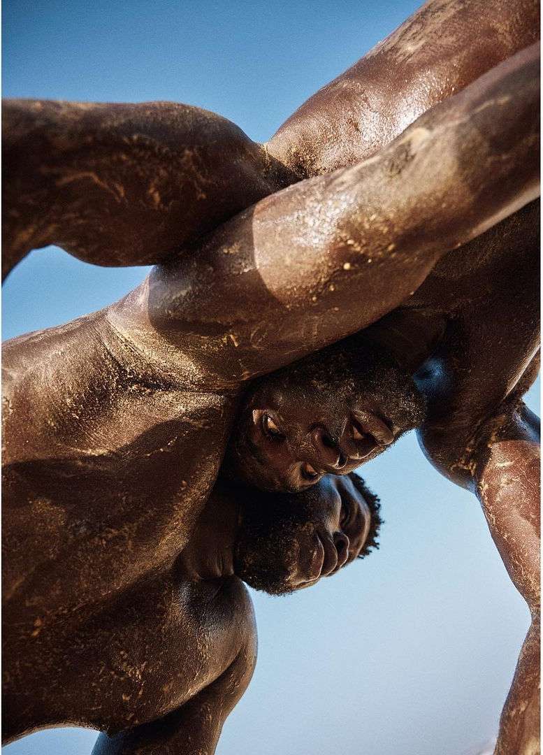 Two dark-skinned adult men in a close wrestling match, bodies entwined and glistening with sweat, set against a clear light blue sky background.