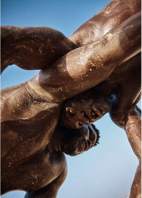 Two dark-skinned adult men in a close wrestling match, bodies entwined and glistening with sweat, set against a clear light blue sky background.