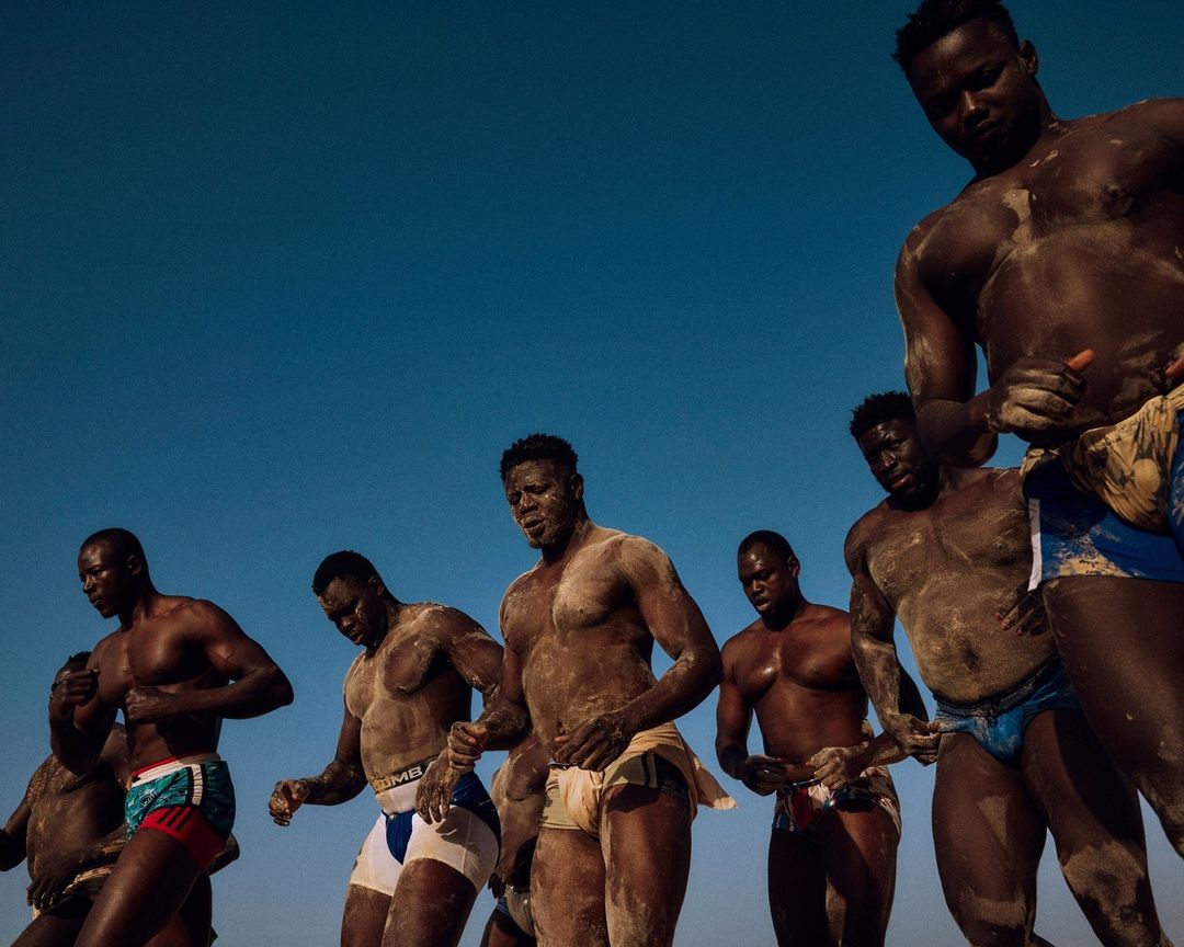 Seven muscular Senegalese wrestlers covered in sand stand in a line against a clear blue sky, preparing for a traditional wrestling match.
