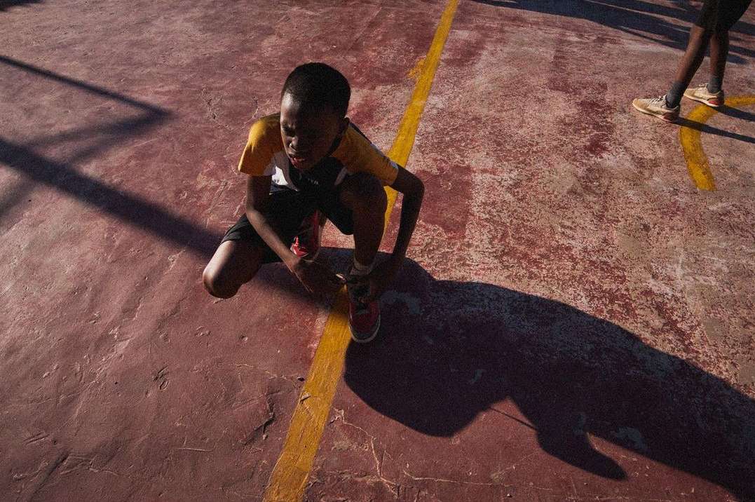 A child tying shoelaces on a worn outdoor court with yellow line, capturing a moment of focus and determination in natural sunlight.