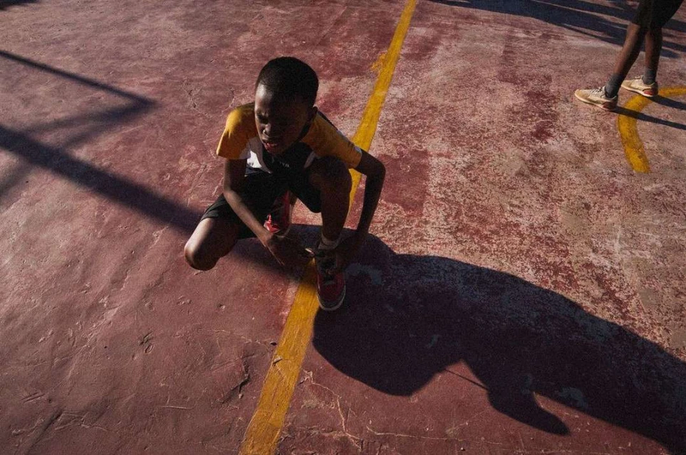 A child tying shoelaces on a worn outdoor court with yellow line, capturing a moment of focus and determination in natural sunlight.