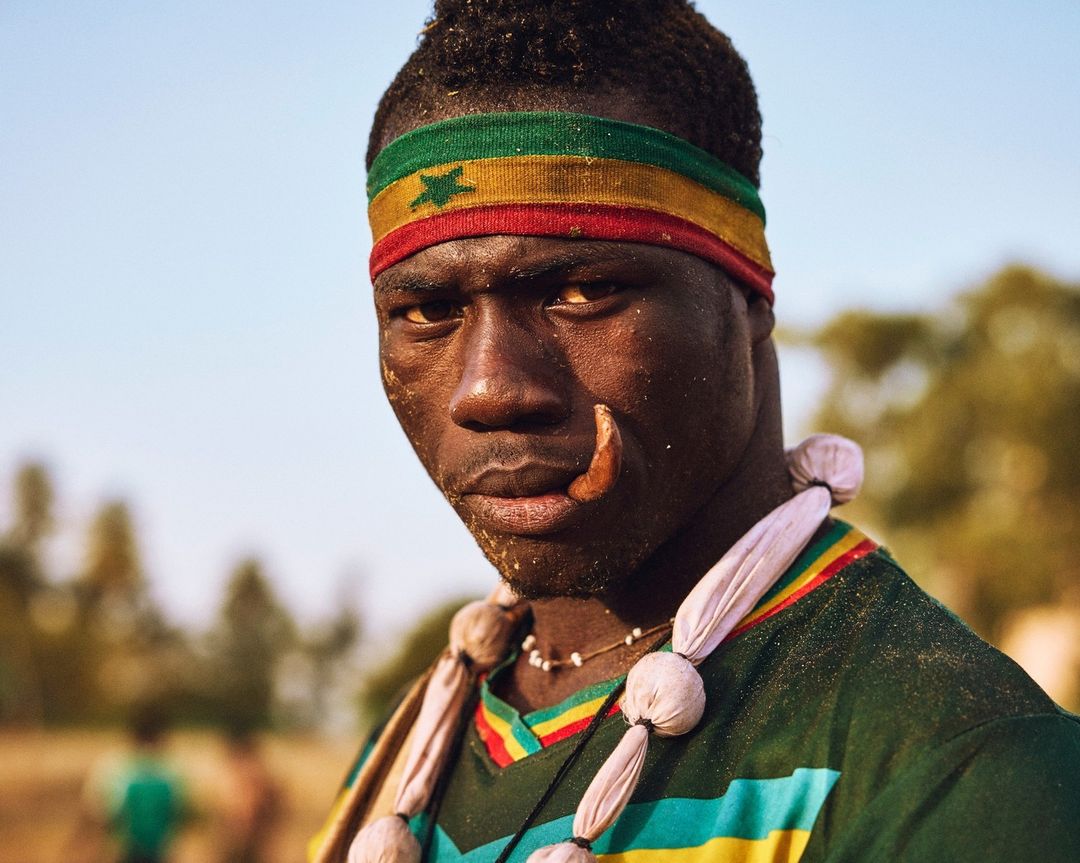 Portrait of a dark-skinned man with a Ghana flag headband, face jewelry, and a necklace, posing outdoors in a close-up shot on a sunny day.