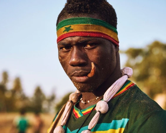 Portrait of a dark-skinned man with a Ghana flag headband, face jewelry, and a necklace, posing outdoors in a close-up shot on a sunny day.