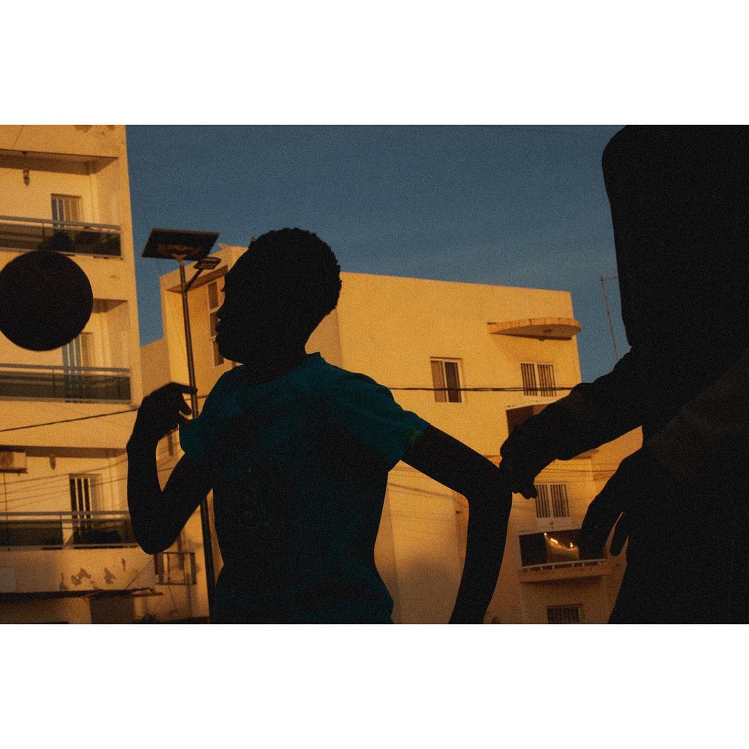 Two silhouetted children playing with a basketball on a rooftop at dusk with apartment buildings visible in the warm-toned background.