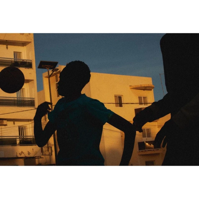 Two silhouetted children playing with a basketball on a rooftop at dusk with apartment buildings visible in the warm-toned background.