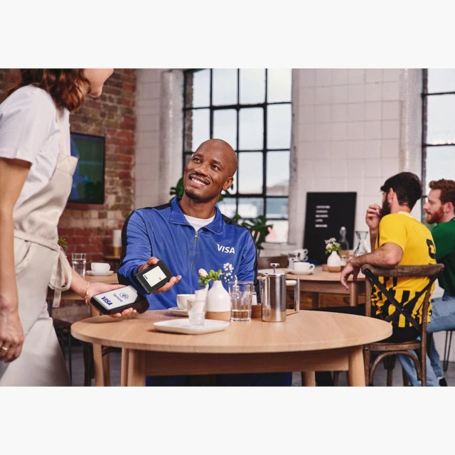 Man in blue jacket pays with contactless card at cafe, held by server; other customers in background. Visa logo visible on jacket and card.